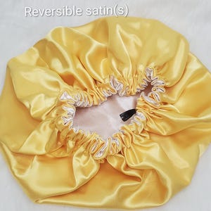 May include: A reversible satin bonnet featuring a bright yellow exterior and a pale cream interior. The hair accessory has a gathered elastic edge for a secure fit and includes the text Reversible satin(s) printed at the top of the image.