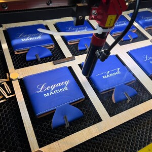 May include: A laser engraver is working on a set of blue can coolers with the text "Legacy Marine" printed on them. The coolers are arranged in a grid pattern on a wooden surface.