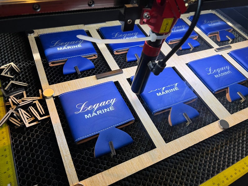 May include: A laser engraver is working on a set of blue can coolers with the text "Legacy Marine" printed on them. The coolers are arranged in a grid pattern on a wooden surface.