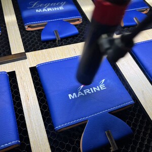 May include: Close-up of a blue leather can cooler with white embroidery that reads "MARINE". The cooler is being engraved with a laser.