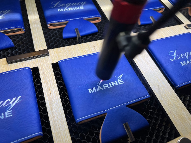 May include: Close-up of a blue leather can cooler with white embroidery that reads "MARINE". The cooler is being engraved with a laser.