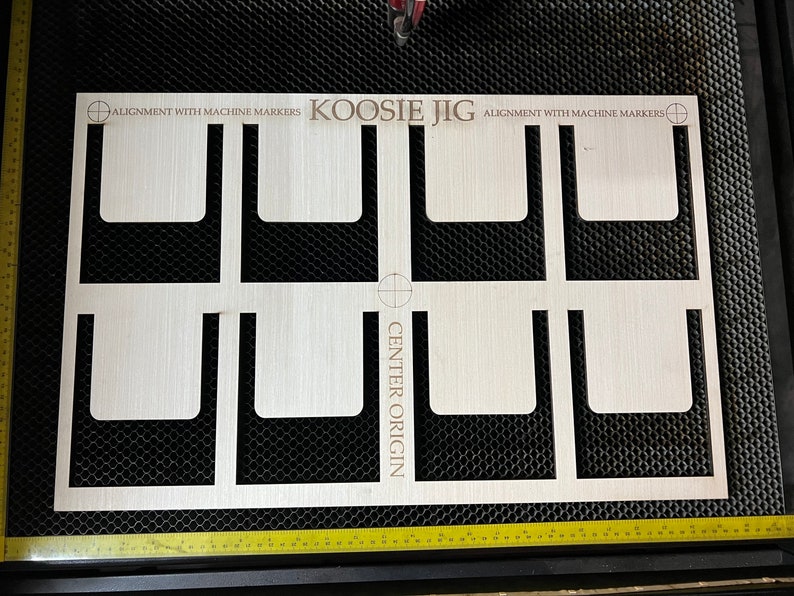 May include: A wooden jig with eight cutouts for aligning objects. The jig has the text "KOOSIE JIG" and "CENTER ORIGIN" printed on it. The text "ALIGNMENT WITH MACHINE MARKERS" is printed twice on the jig.