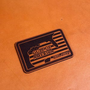 Puede incluir: Un parche de cuero marrón con un contorno negro y un diseño de cuerda. El parche presenta una silueta del estado de Florida con un patrón de bandera estadounidense y el texto "GULF OF AMERICA EST. 2025".