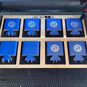 May include: Eight blue neoprene can coolers with a white logo that says "The Williamsburg Kitchen" and a palm tree graphic. The coolers are arranged in two rows of four on a wooden surface with a honeycomb pattern.