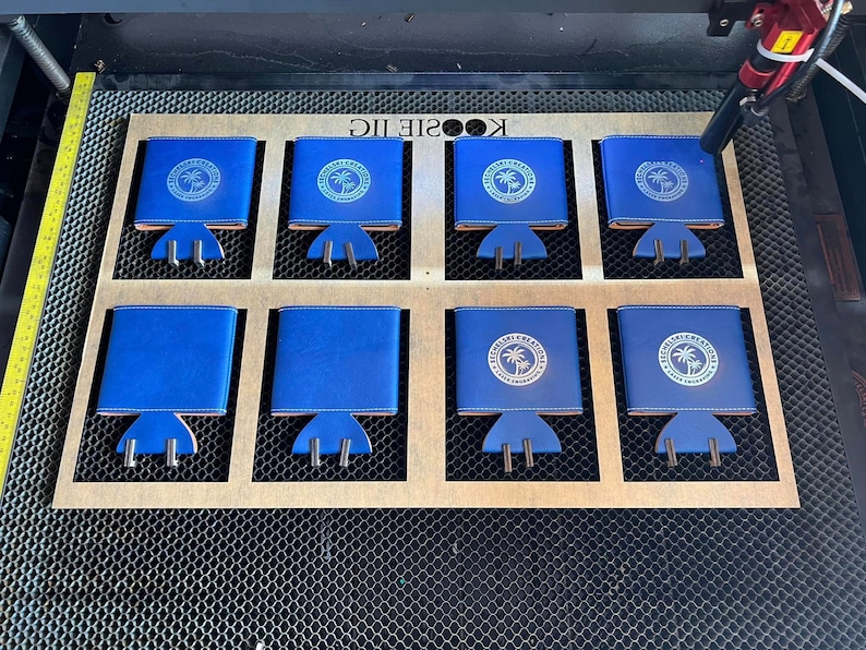 May include: Eight blue neoprene can coolers with a white logo that says "The Williamsburg Kitchen" and a palm tree graphic. The coolers are arranged in two rows of four on a wooden surface with a honeycomb pattern.
