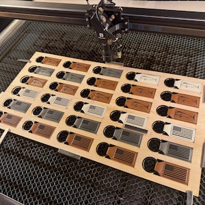 May include: A wooden board with multiple cutouts, holding various leather keychains. The keychains feature designs like the American flag and a fish. The laser cutter is visible above the board, indicating the production process.