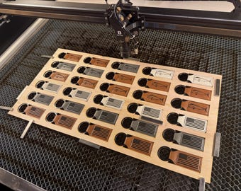 Puede incluir: Una tabla de madera con múltiples recortes, que contiene varios llaveros de cuero. Los llaveros presentan diseños como la bandera estadounidense y un pez. El cortador láser es visible encima de la tabla.