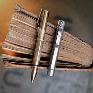 Könnte beinhalten: Zwei Metallstifte, einer goldfarben und einer silberfarben, liegen auf den gealterten Seiten eines offenen Buches. Der goldfarbene Stift hat einen Clip und einen strukturierten Griff. Der silberne Stift hat ein Fuchslogo auf dem Clip.