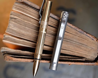 May include: Two metal pens, one gold-toned and one silver, rest against the aged pages of an open book. The gold pen has a clip and textured grip. The silver pen features a fox logo on its clip. The book's pages are yellowed with age.