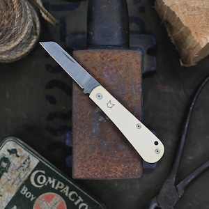 May include: A small, folding pocket knife with a cream-colored handle and a silver blade. The handle has a small fox emblem and is secured with three small screws. The knife is resting on a rusty metal surface.