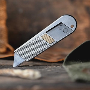 May include: A silver-toned utility knife with a retractable blade, featuring a textured diamond-patterned grip, a small rectangular gold-coloured accent, and two circular indentations near the rounded end which includes a small lanyard hole.