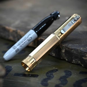 Puede incluir: Un rotulador Sharpie negro junto a un bolígrafo dorado. El bolígrafo tiene un clip plateado con un pequeño diseño y un extremo texturizado. El Sharpie tiene una tapa negra y la palabra "Sharpie" impresa en el cuerpo blanco.