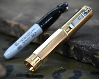 May include: A black Sharpie marker next to a gold-colored pen. The pen has a silver clip with a small design and a textured end. The Sharpie has a black cap and the word "Sharpie" printed on the white barrel.