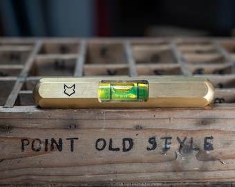 May include: A brass level with a green bubble and a fox logo. The level is resting on a wooden box with the text "POINT OLD STYLE" printed on it.