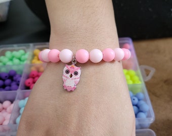 May include: Pink and white beaded bracelet with an owl charm. The owl is pink with white eyes and a white beak.