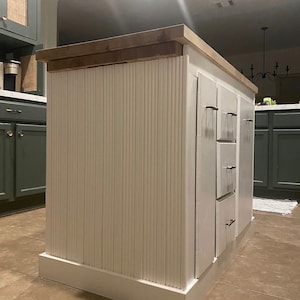 Puede incluir: Isla de cocina blanca con encimera de madera y paneles verticales blancos. La isla tiene tres cajones con tiradores plateados.