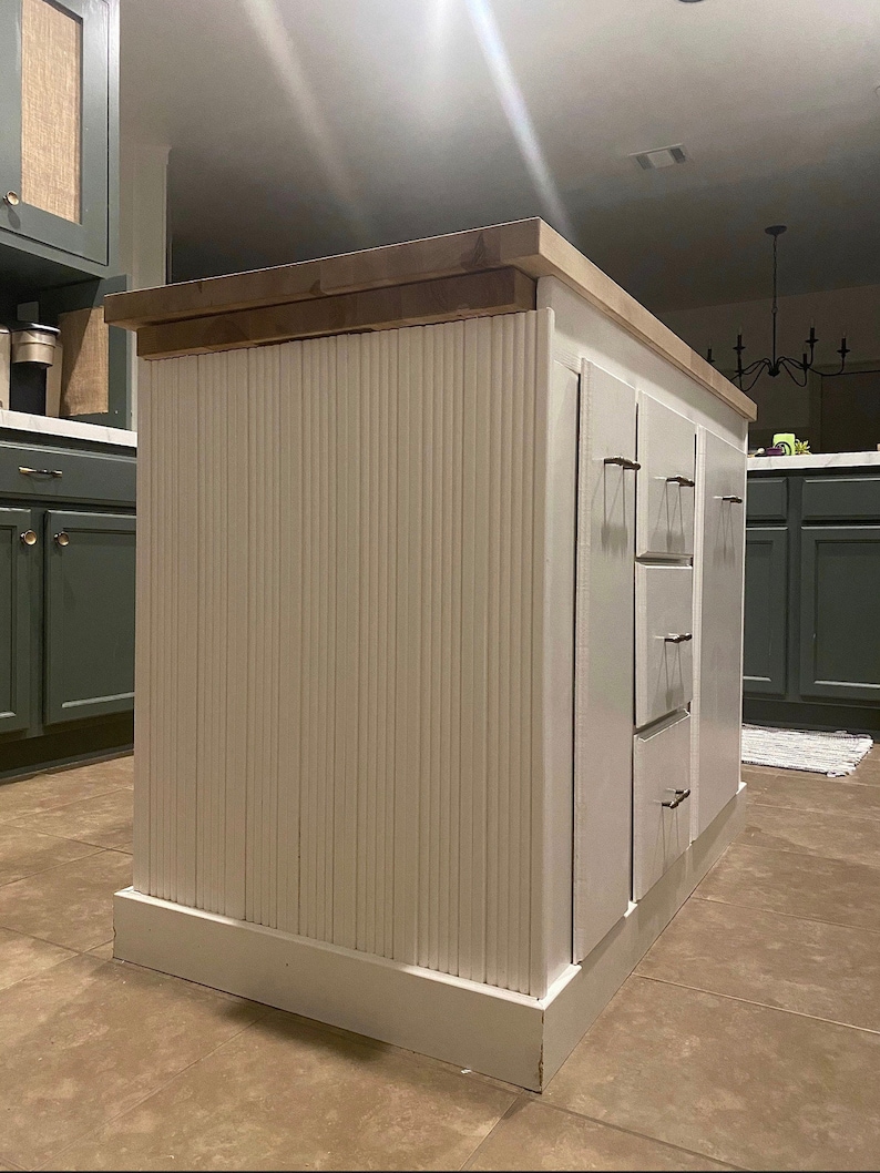 Puede incluir: Isla de cocina blanca con encimera de madera y paneles verticales blancos. La isla tiene tres cajones con tiradores plateados.