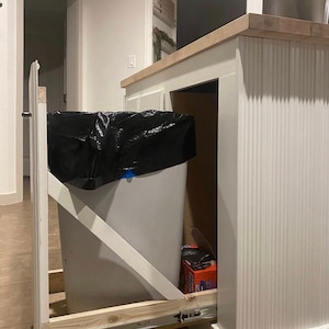 Puede incluir: Un cubo de basura blanco extra&iacute;ble con una bolsa de basura negra en su interior, montado debajo de una encimera de cocina. El cubo de basura est&aacute; sobre una v&iacute;a deslizante y tiene un marco de madera.