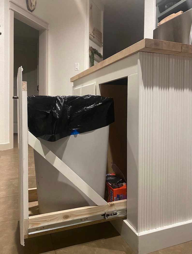 Puede incluir: Un cubo de basura blanco extra&iacute;ble con una bolsa de basura negra en su interior, montado debajo de una encimera de cocina. El cubo de basura est&aacute; sobre una v&iacute;a deslizante y tiene un marco de madera.