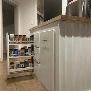 Puede incluir: Armario de cocina blanco con un estante de especias extra&iacute;ble. El estante de especias est&aacute; lleno de varios frascos de especias. El armario tiene un acabado blanco texturizado y una z&oacute;calo blanco.