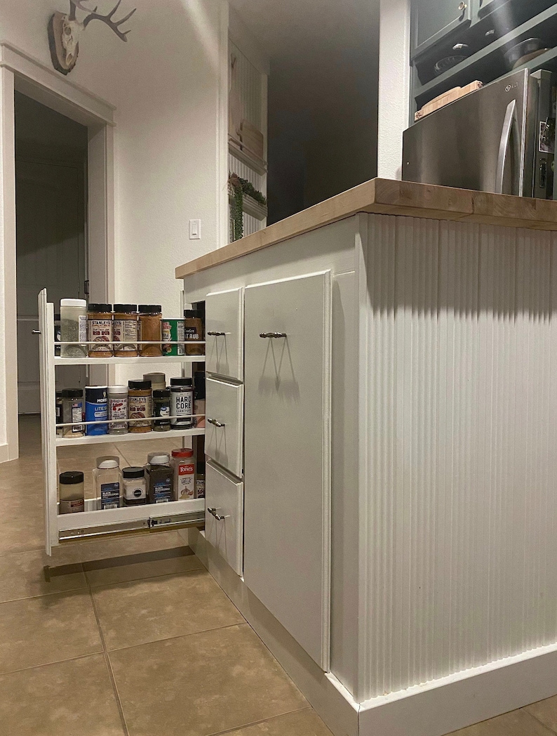 Puede incluir: Armario de cocina blanco con un estante de especias extra&iacute;ble. El estante de especias est&aacute; lleno de varios frascos de especias. El armario tiene un acabado blanco texturizado y una z&oacute;calo blanco.