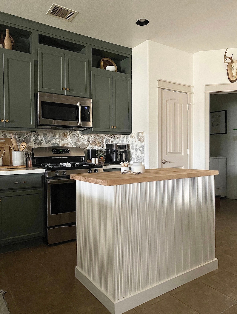 Puede incluir: Una isla de cocina con una encimera de madera y una base de metal corrugado blanco. La isla est&aacute; frente a una cocina verde con electrodom&eacute;sticos de acero inoxidable.