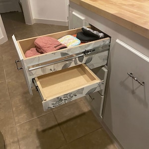 Puede incluir: Dos cajones abiertos en un armario de cocina blanco. El caj&oacute;n superior est&aacute; parcialmente abierto y contiene un par de guantes de horno rosas, un posavasos azul y blanco y un guante de horno negro. El caj&oacute;n inferior est&aacute; completamente abierto y vac&iacute;o. Los cajones tienen un patr&oacute;n floral en la parte delantera.