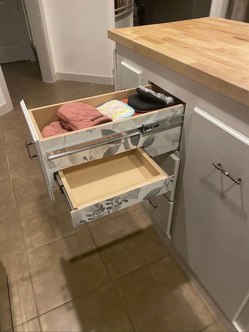 Puede incluir: Dos cajones abiertos en un armario de cocina blanco. El caj&oacute;n superior est&aacute; parcialmente abierto y contiene un par de guantes de horno rosas, un posavasos azul y blanco y un guante de horno negro. El caj&oacute;n inferior est&aacute; completamente abierto y vac&iacute;o. Los cajones tienen un patr&oacute;n floral en la parte delantera.