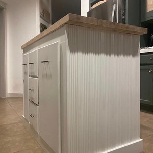 Puede incluir: Isla de cocina blanca con encimera de madera y panel frontal acanalado. La isla tiene tres cajones con herrajes de n&iacute;quel cepillado.