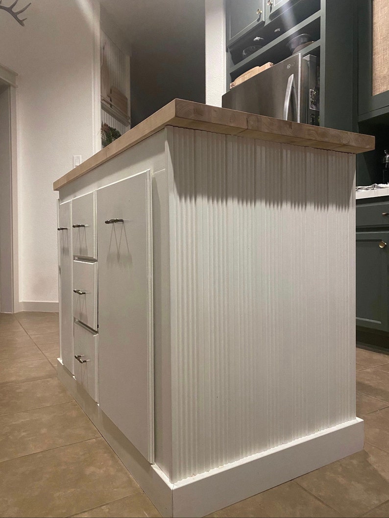 Puede incluir: Isla de cocina blanca con encimera de madera y panel frontal acanalado. La isla tiene tres cajones con herrajes de n&iacute;quel cepillado.