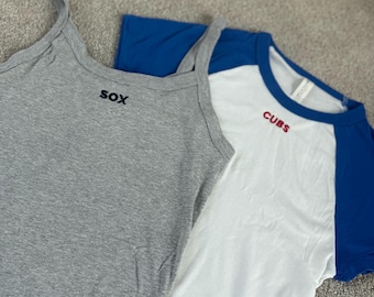May include: Two athletic tops. A grey vest top with "SOX" embroidered in black, and a white and blue baseball tee with "CUBS" embroidered in red. The tops are made of soft fabric.