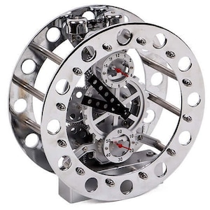 Può includere: Un orologio meccanico color argento con ingranaggi a vista e due quadranti circolari. L'orologio ha un design unico con una cornice a forma di ruota e una base rettangolare. Le lancette dell'orologio sono rosse.