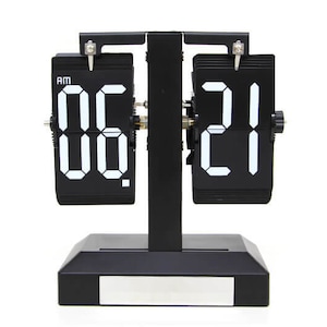 Può includere: Un orologio digitale a scatto in bianco e nero che mostra l'ora 06:21 AM. L'orologio ha una base rettangolare e un supporto centrale che tiene i display dell'ora. I numeri sono bianchi su sfondo nero.