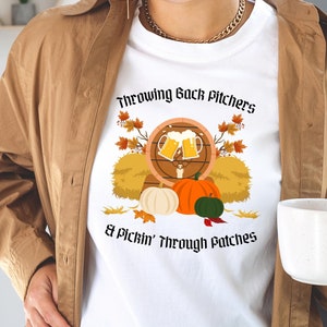 May include: White t-shirt with the text "Throwing Back Pitchers & Pickin' Through Patches" in a fall-themed design. The graphic includes beer mugs, pumpkins, and autumn leaves. The person is wearing a brown jacket.