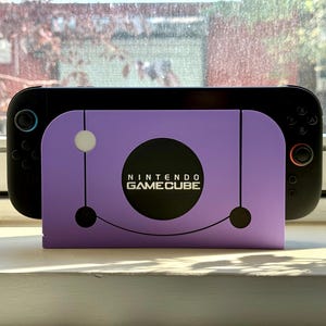 May include: A black handheld gaming device with a purple Nintendo GameCube-themed skin. The skin features the GameCube logo and design. The device is resting on a white surface near a window.