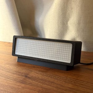 Könnte beinhalten: Ein schwarzes rechteckiges LED-Display mit einem weißen Punktmatrix-Bildschirm. Das Display hat einen strukturierten schwarzen Rahmen und steht auf einem schwarzen Sockel. Ein schwarzes Netzkabel erstreckt sich von der Rückseite. Das Gerät steht auf einer Holzoberfläche.