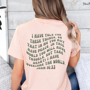 May include: A pink t-shirt with a green text graphic that reads "I have told you these things, so that in me you may have peace. In this world you will have trouble. But take heart! I have overcome the world. John 16:33"