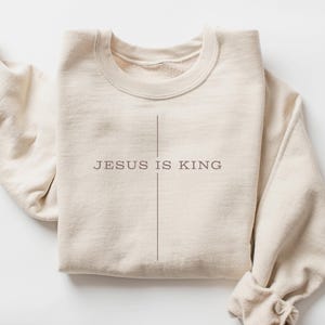 May include: A beige sweatshirt with the text "JESUS IS KING" printed on the front. The text is centered and is in a simple, modern font.