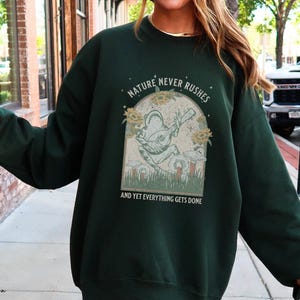 May include: Forest green sweatshirt with a graphic of a frog playing a guitar and the text "Nature never rushes and yet everything gets done".