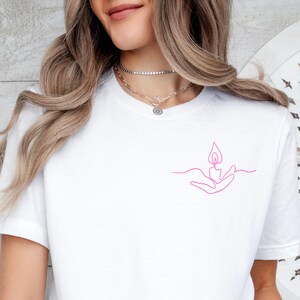 May include: White t-shirt with a pink line drawing of a hand holding a flame.