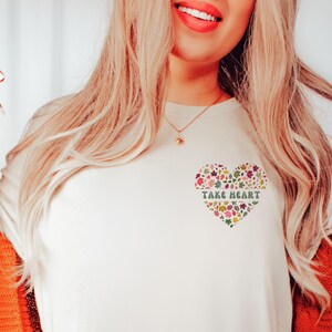 May include: White t-shirt with a floral heart design and the text "Take Heart" in green letters.