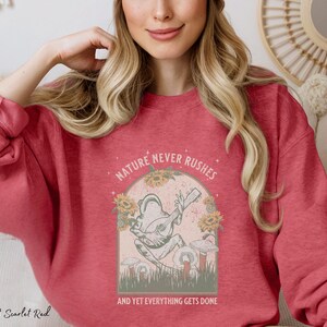 May include: A heather sport scarlet red sweatshirt with a graphic of a frog playing a guitar. The graphic is surrounded by flowers and the text "Nature never rushes and yet everything gets done".