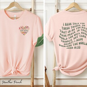 May include: Pink t-shirt with a floral heart design on the front and a quote from John 16:33 on the back. The quote reads "I have told you these things, so that in me you may have peace. In this world you will have trouble. But take heart! I have overcome the world."