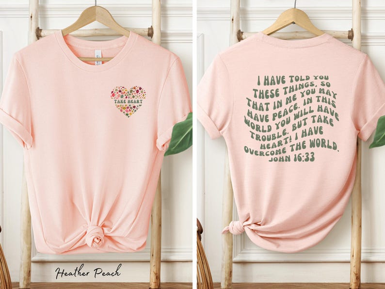 May include: Pink t-shirt with a floral heart design on the front and a quote from John 16:33 on the back. The quote reads "I have told you these things, so that in me you may have peace. In this world you will have trouble. But take heart! I have overcome the world."