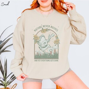 May include: A beige sweatshirt with a graphic of a frog playing a guitar in a forest setting. The text "Nature Never Rushes And Yet Everything Gets Done" is printed below the graphic.