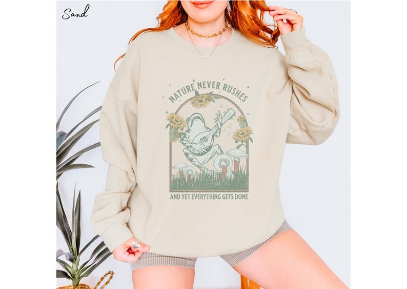 May include: A beige sweatshirt with a graphic of a frog playing a guitar in a forest setting. The text "Nature Never Rushes And Yet Everything Gets Done" is printed below the graphic.