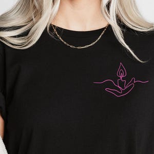 May include: A black t-shirt with a pink line drawing of a hand holding a candle flame.
