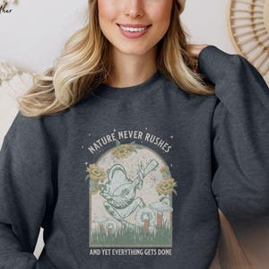 May include: A dark heather grey sweatshirt with a graphic of a frog playing a banjo. The graphic is surrounded by flowers and mushrooms. The text "NATURE NEVER RUSHES AND YET EVERYTHING GETS DONE" is printed below the graphic.