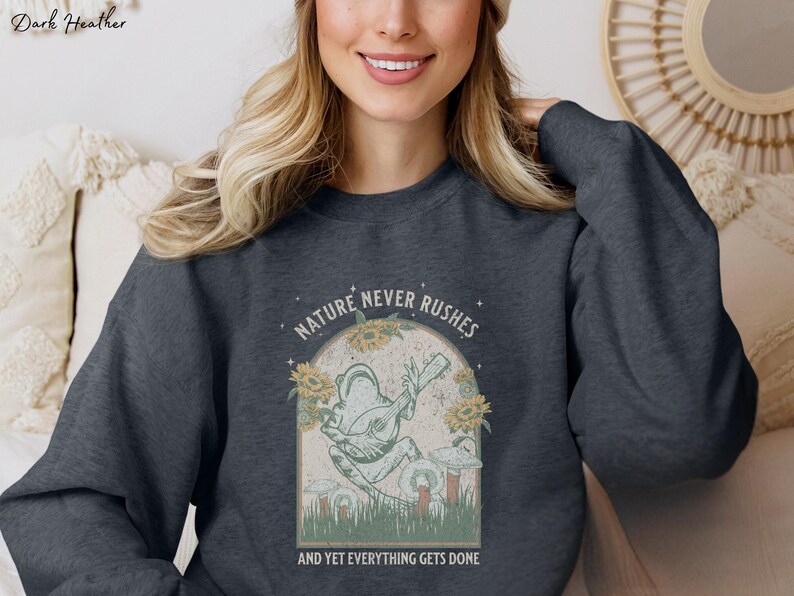 May include: A dark heather grey sweatshirt with a graphic of a frog playing a banjo. The graphic is surrounded by flowers and mushrooms. The text "NATURE NEVER RUSHES AND YET EVERYTHING GETS DONE" is printed below the graphic.