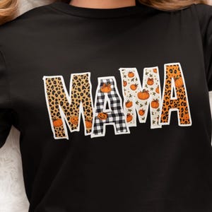 Puede incluir: Camiseta negra de manga larga con la palabra "MAMA" en una fuente decorativa. Las letras tienen un estampado de leopardo, calabaza y cuadros de búfalo. La camiseta probablemente está hecha de un material suave y cómodo.